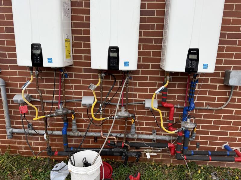 Three tankless water heaters installed side by side on a brick wall with gas and water piping